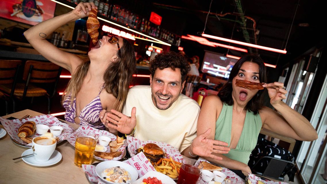 A WeRoad group trip of three people having a fun brunch, making funny faces with a croissant and bacon in a restaurant.