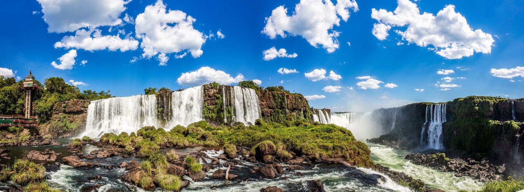 Une vue panoramique d'un vaste système de chutes d'eau se déversant sur des falaises rocheuses recouvertes de végétation verdoyante sous un ciel bleu clair et nuageux.