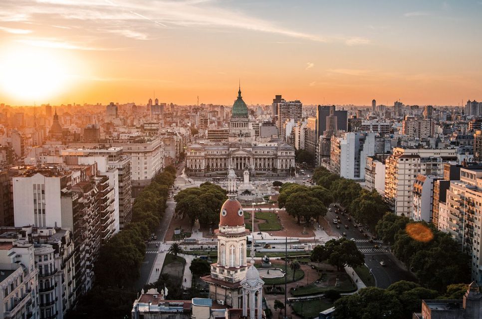 An aerial view of a dense cityscape centered on a large building with a green dome and a plaza at sunset.