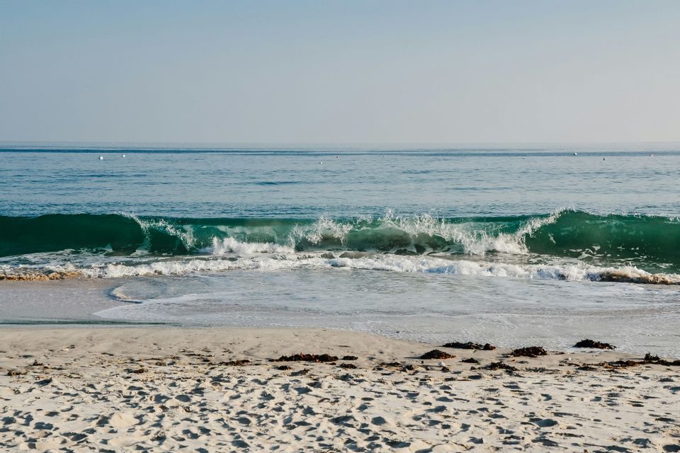 Une vague turquoise avec de l'écume blanche se brise sur une plage de sable sous un ciel clair.