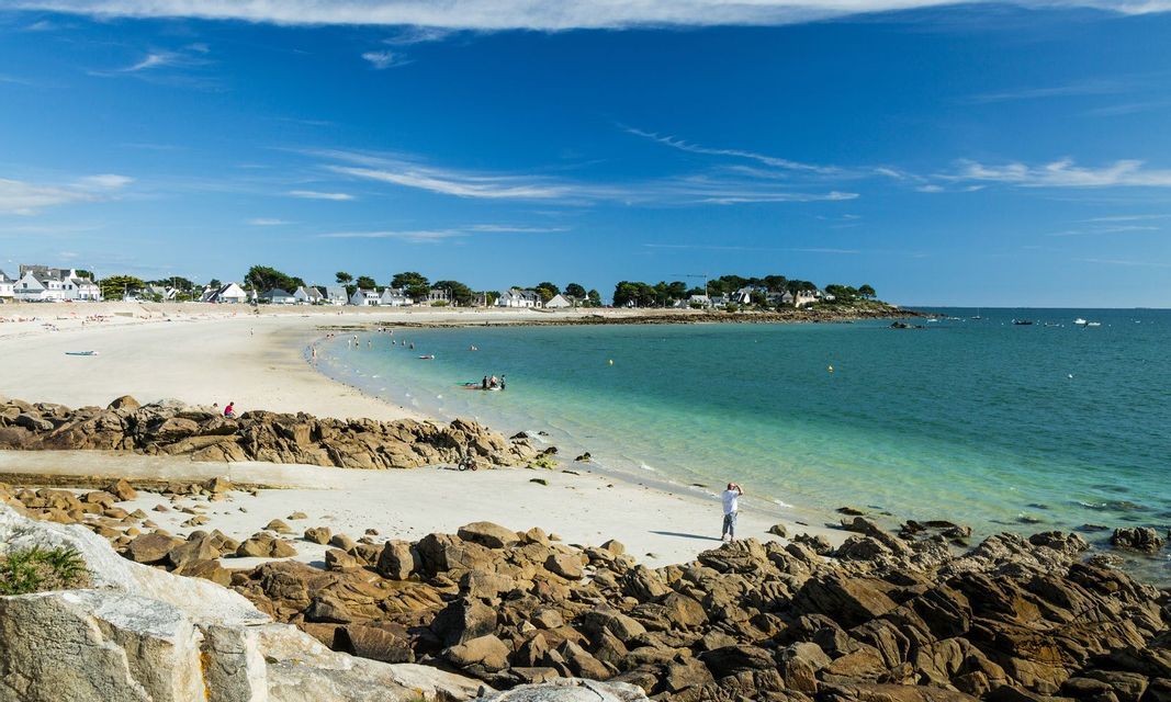 Une côte rocheuse surplombe une plage de sable blanc et une crique turquoise avec un petit village en arrière-plan sous un ciel bleu.