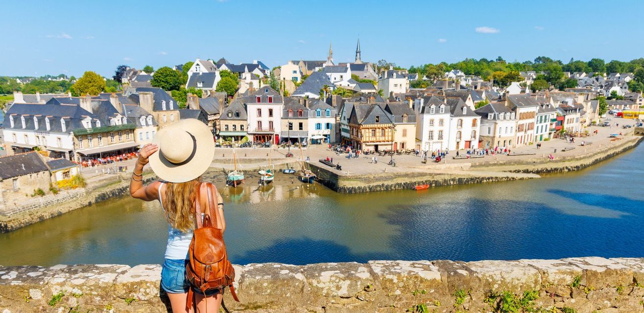 Une femme coiffée d'un chapeau de paille et portant un sac à dos contemple depuis un mur de pierre une ville portuaire européenne avec des bateaux.
