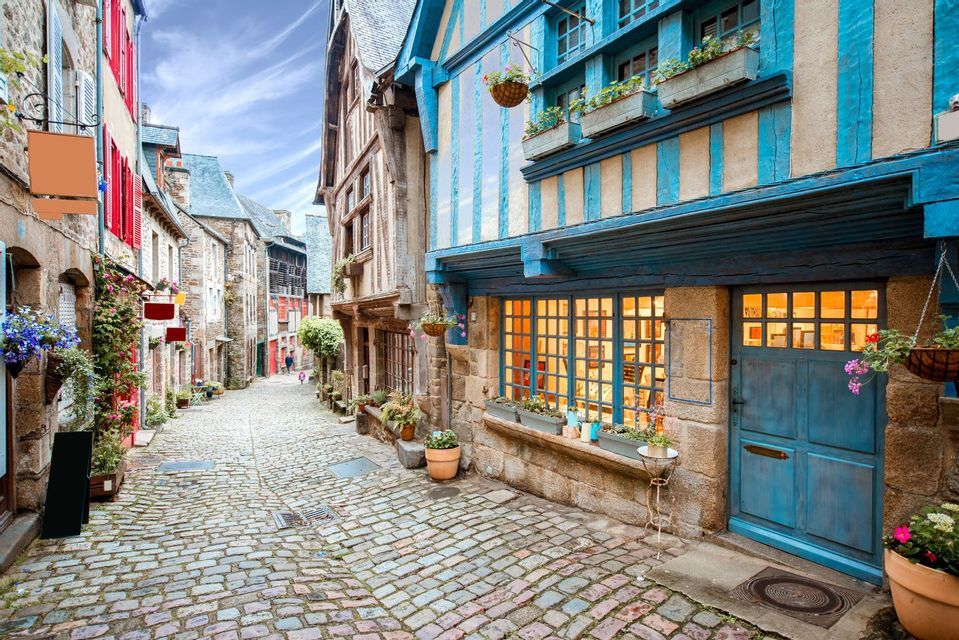 Une rue étroite pavée serpente devant des maisons historiques à colombages avec des détails bleus et rouges sous un ciel partiellement nuageux.