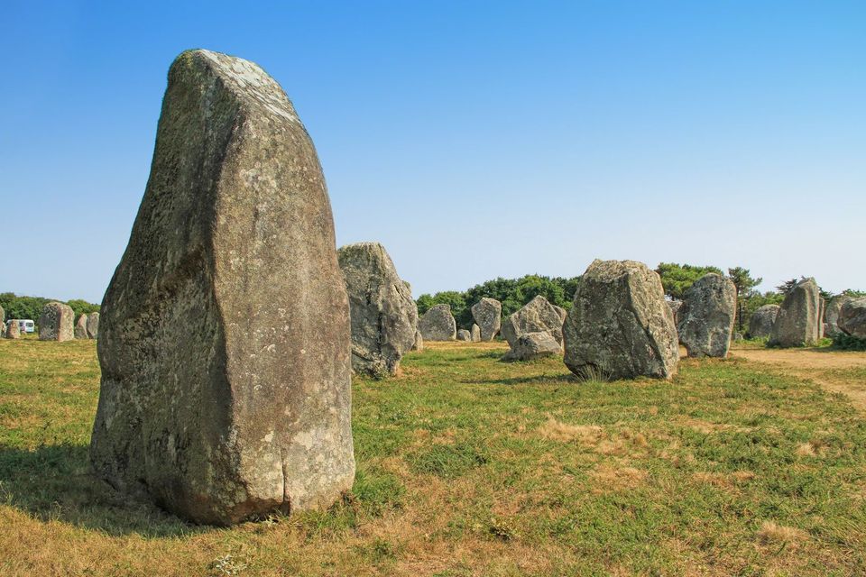 Un grand mégalithe se dresse au premier plan d'un champ de menhirs sous un ciel d'un bleu éclatant.