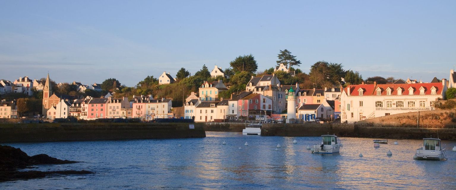Un village côtier avec des maisons colorées et un petit phare est perché sur une colline surplombant un port avec plusieurs bateaux.