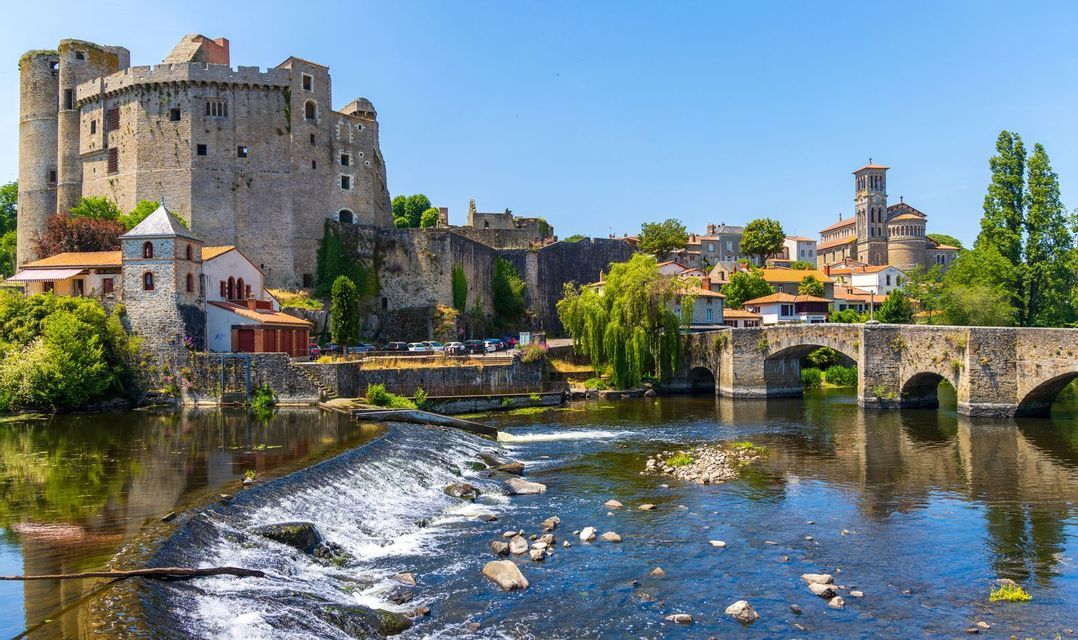 Un château médiéval en pierre et un pont en arc enjambent une rivière avec un petit barrage dans une ville historique sous un ciel bleu clair.