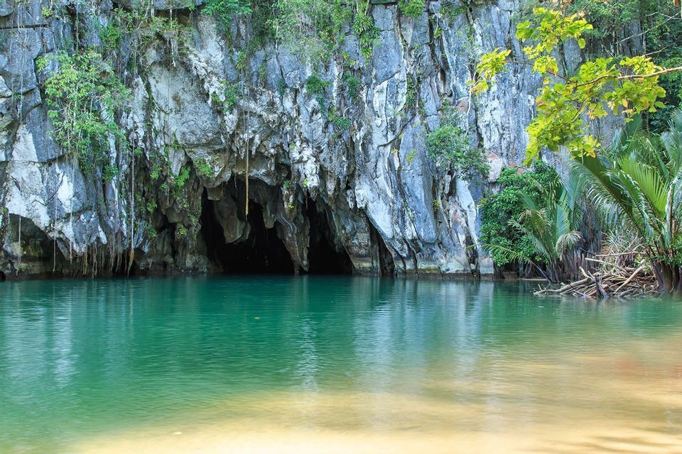 Una oscura entrada a una cueva al pie de un acantilado rocoso cubierto de vegetación, que da a un cuerpo de agua tranquila de color verde esmeralda con una orilla arenosa.