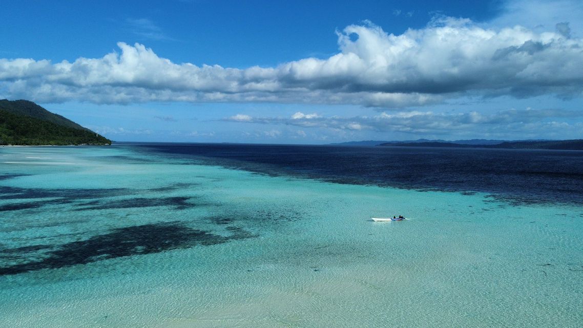 Vista aerea di una piccola barca su acqua turchese poco profonda accanto al mare blu profondo, con un'isola tropicale sullo sfondo.