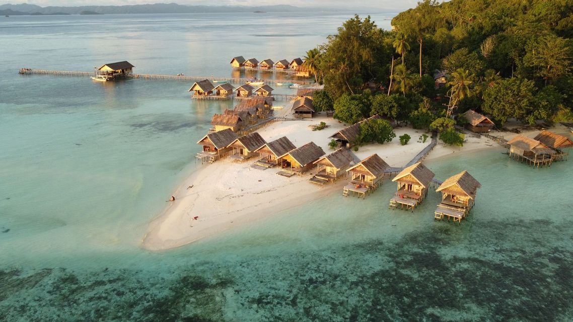 Un villaggio di bungalow sull'acqua con tetti di paglia su una spiaggia di sabbia bianca, accanto a un'isola tropicale lussureggiante e un mare turchese.