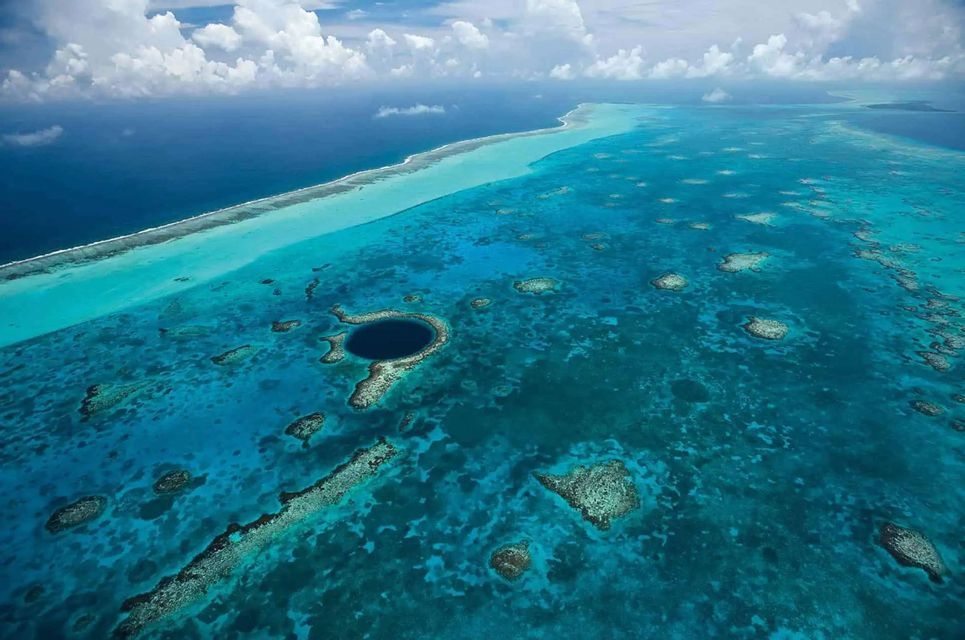 Aerial view of a large, dark blue marine sinkhole surrounded by turquoise water and coral reefs under a partly cloudy sky.