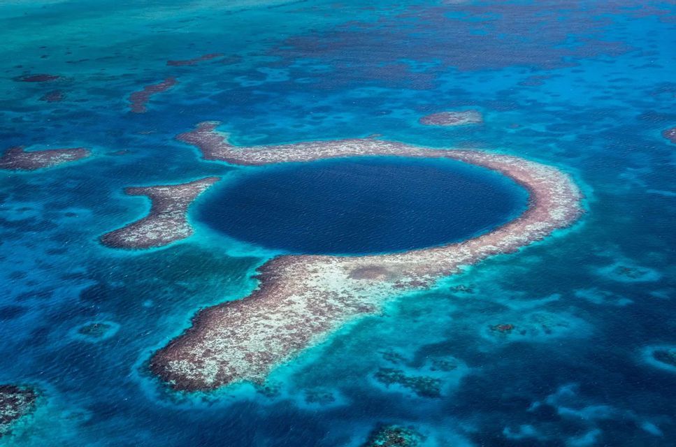 An aerial shot of a large, dark blue marine sinkhole encircled by a coral reef atoll in turquoise waters.