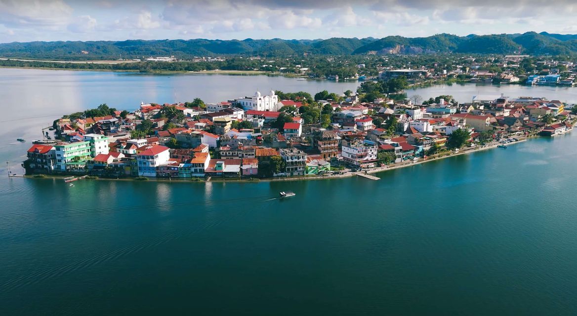 An aerial view of a colorful town on a peninsula in a large blue lake, with green hills visible in the background.