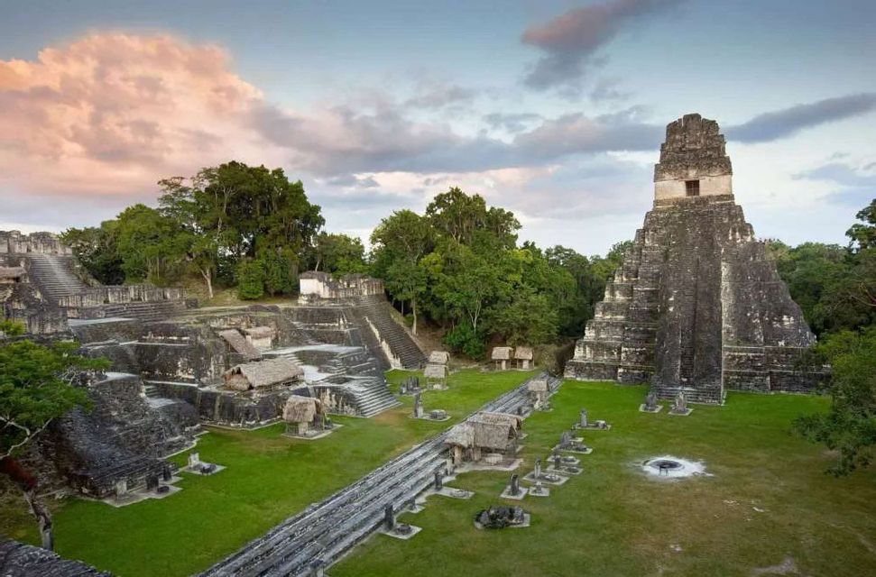 An expansive view of ancient stone pyramids and ruins surrounded by dense green jungle under a cloudy sky at dusk.