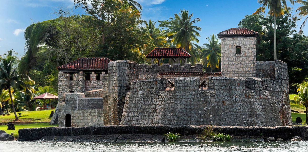 A stone fortress with tiled roofs and cannons on its ramparts stands on a shore surrounded by lush palm trees and greenery.