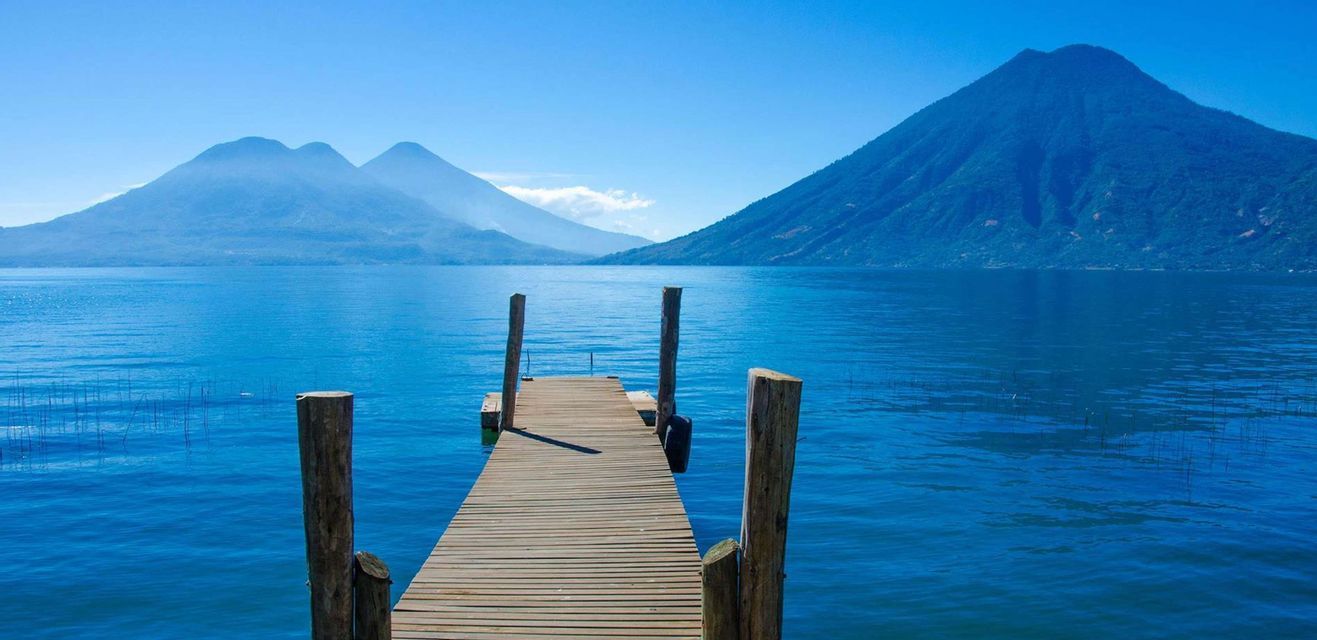 A wooden pier extends over a calm blue lake, with large mountains visible in the distance under a clear sky.