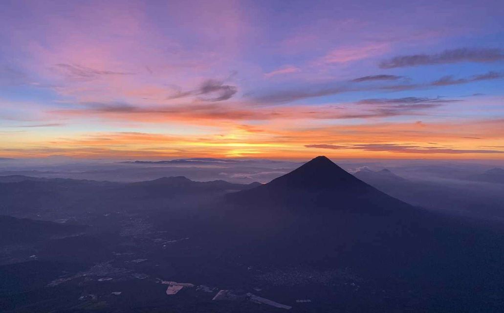 A volcanic cone rises above a misty valley under a vibrant orange and purple sunrise sky.