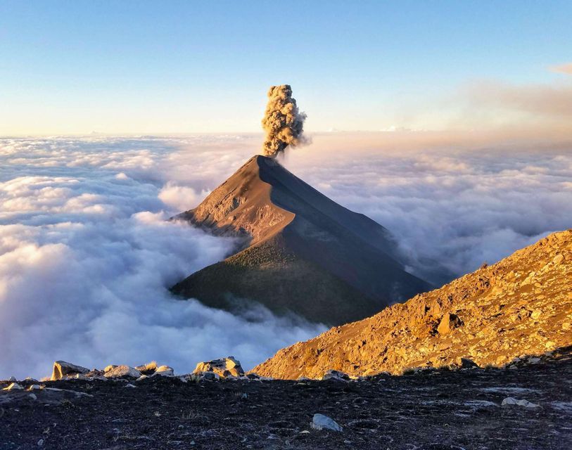 A volcano erupts with a plume of ash, its peak rising above a sea of clouds under a clear sky at sunrise.