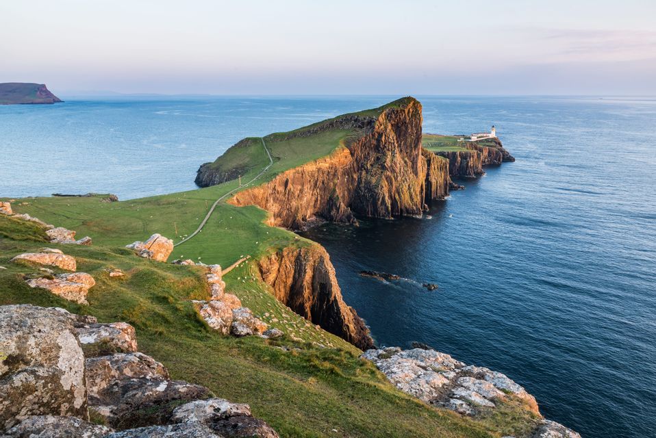 Un faro blanco se erige en una península verde y acantilada que se adentra en un mar azul y tranquilo bajo un cielo despejado.