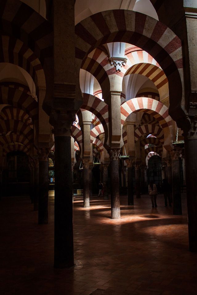 Interno di una sala storica con numerose colonne che sorreggono archi a strisce rosse e bianche, illuminato da fasci di luce.