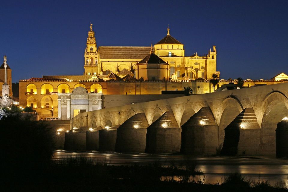Un ponte in pietra illuminato a più arcate attraversa un fiume di notte, con un grande edificio storico sullo sfondo.