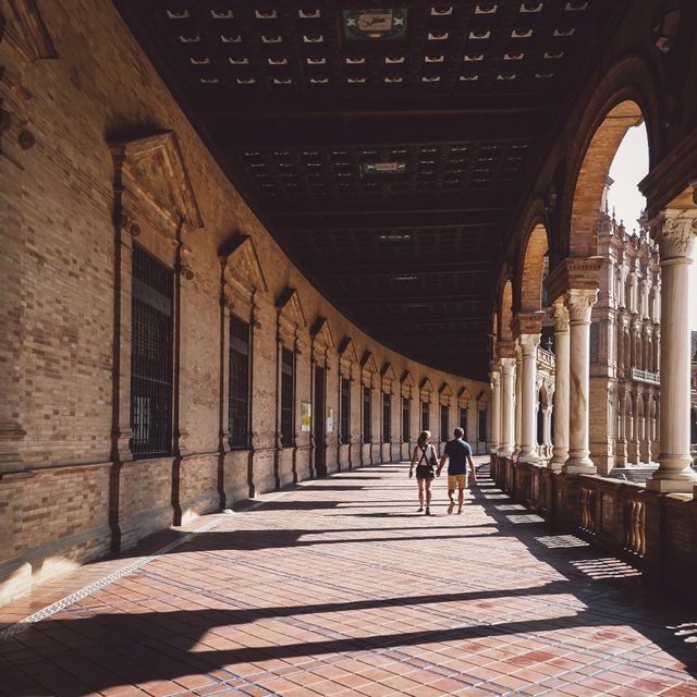 Una coppia cammina mano nella mano lungo un lungo portico curvo con muri di mattoni e archi, creando lunghe ombre.