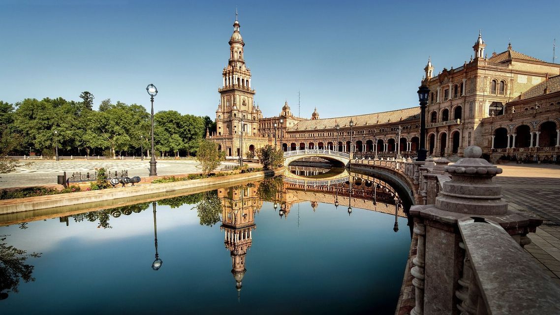 Un edificio decorato con un'alta torre si riflette nell'acqua calma di un canale, con un ponte che lo attraversa sotto un cielo azzurro limpido.