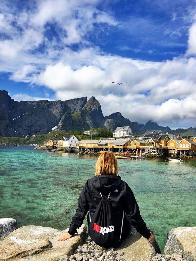 Una persona de espaldas, llevando una mochila WeRoad, se sienta en una roca con vistas a un pueblo de casas amarillas sobre pilotes en agua turquesa, con montañas de fondo.