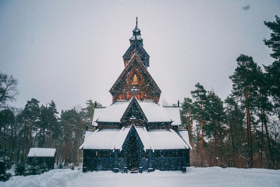 Una tradizionale chiesa a pali in legno si erge in una pineta, i suoi tetti coperti di neve mentre i fiocchi di neve cadono dal cielo.