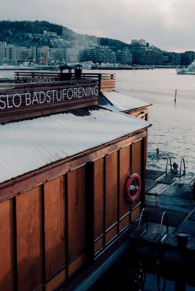 Due persone che fanno il bagno in acqua fredda vicino a una sauna galleggiante in legno con tetto innevato e uno skyline urbano sullo sfondo.