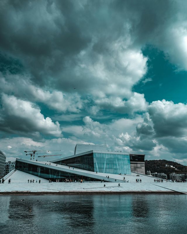 Un moderno edificio con tetto bianco spiovente e pareti di vetro si staglia sull'acqua sotto un cielo drammatico, nuvoloso e color ottanio.