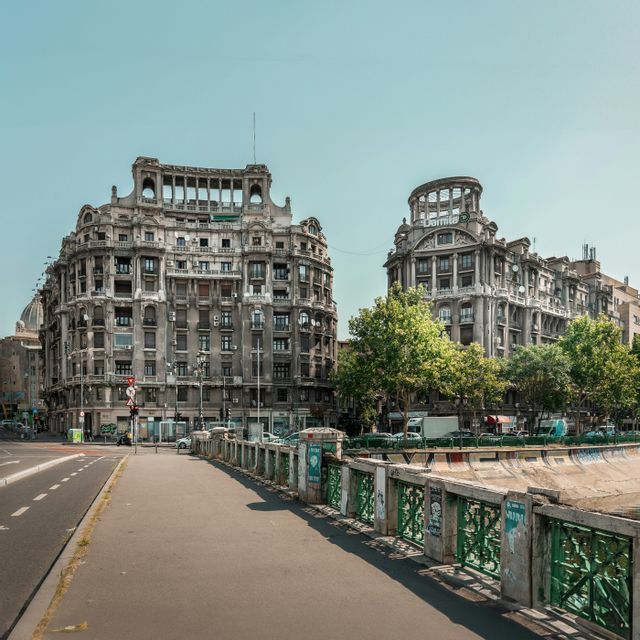 Un marciapiede costeggia un canale fluviale con una ringhiera verde, conducendo verso due grandi edifici storici sotto un cielo limpido.