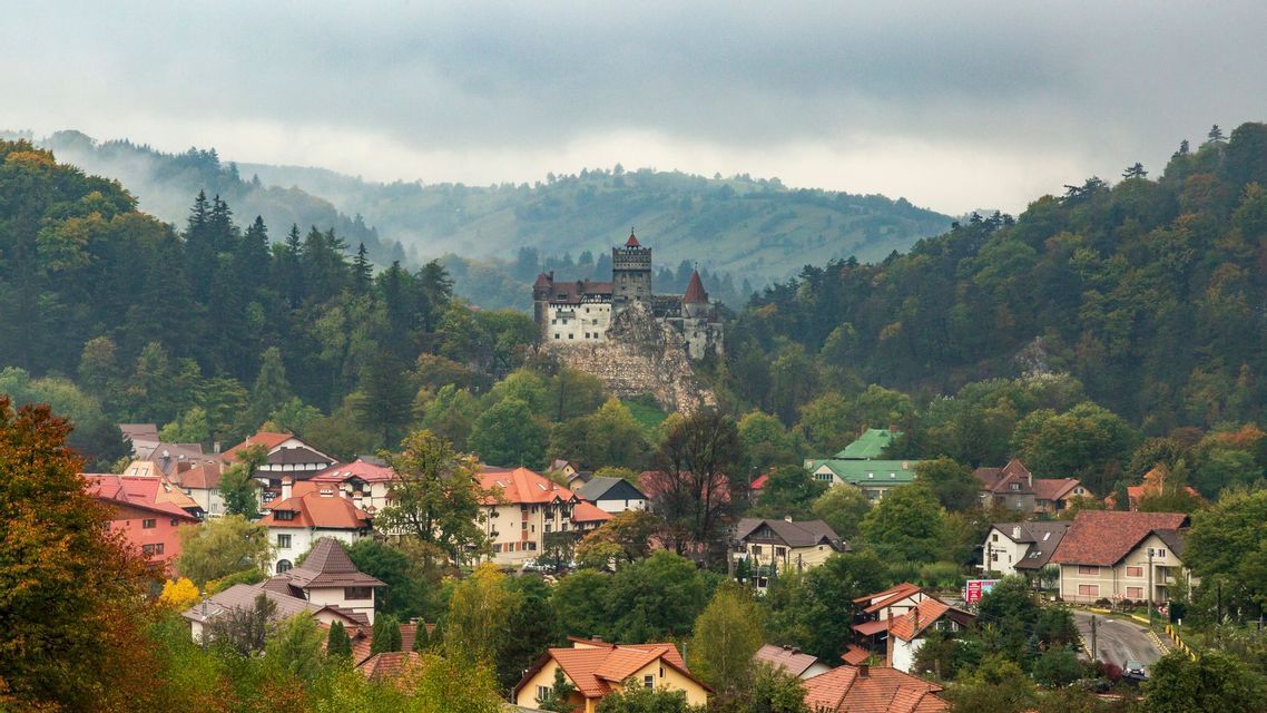 Un castello medievale arroccato su una collina rocciosa che domina un villaggio circondato da una fitta foresta e montagne nebbiose