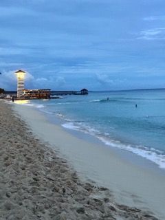 A small, lit lighthouse stands on a sandy beach next to the calm ocean at dusk under a cloudy sky.