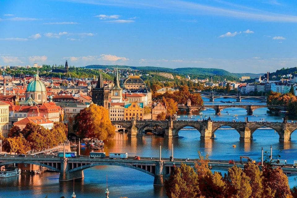 Une vue panoramique d'une ville européenne en automne, avec plusieurs ponts en pierre traversant une large rivière bleue sous un ciel clair.
