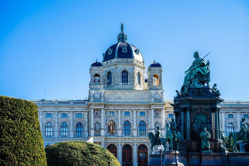 Un bâtiment historique orné, doté d'un grand dôme, se dresse derrière un monument en bronze élaboré sous un ciel bleu clair.