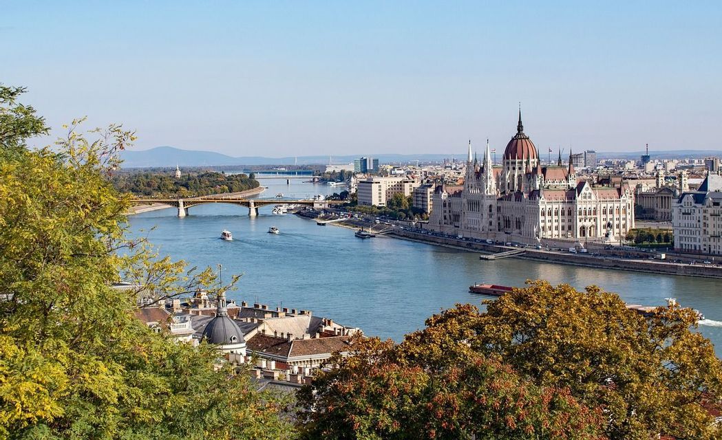 Vue panoramique d'un parlement orné en bord de rivière, avec des bateaux et un pont traversant vers la ville sous un ciel clair.