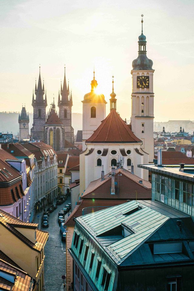 Vue plongeante sur une ville historique avec des flèches gothiques et une tour d'horloge, le soleil brillant derrière les bâtiments.