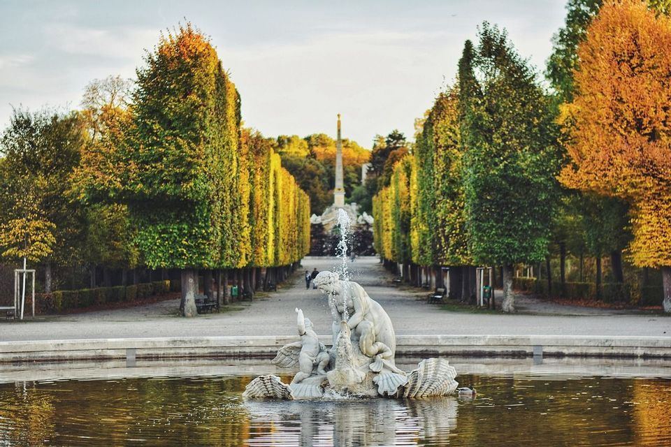 Une fontaine sculpturale se trouve dans un étang au bout d'une longue allée bordée de grands arbres taillés aux couleurs d'automne.