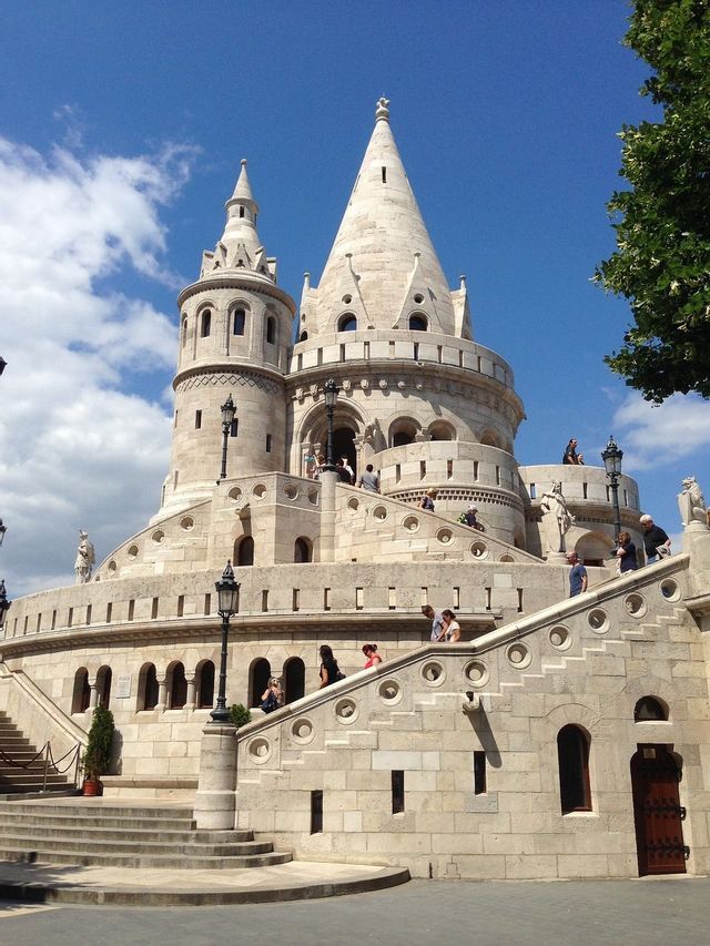 Un bastion en pierre orné avec des tours coniques et de multiples escaliers, exploré par des gens sous un ciel bleu.