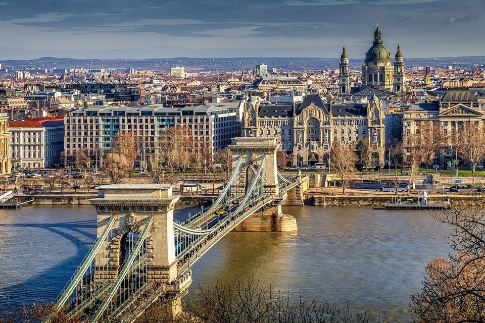 Vue d'un pont suspendu orné traversant une rivière vers un paysage urbain historique avec un bâtiment à dôme proéminent.