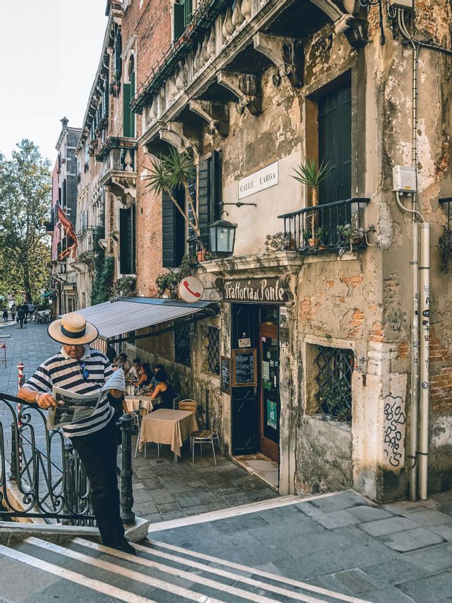 A man in a striped shirt and straw hat reads a newspaper on steps overlooking a trattoria on a narrow street.