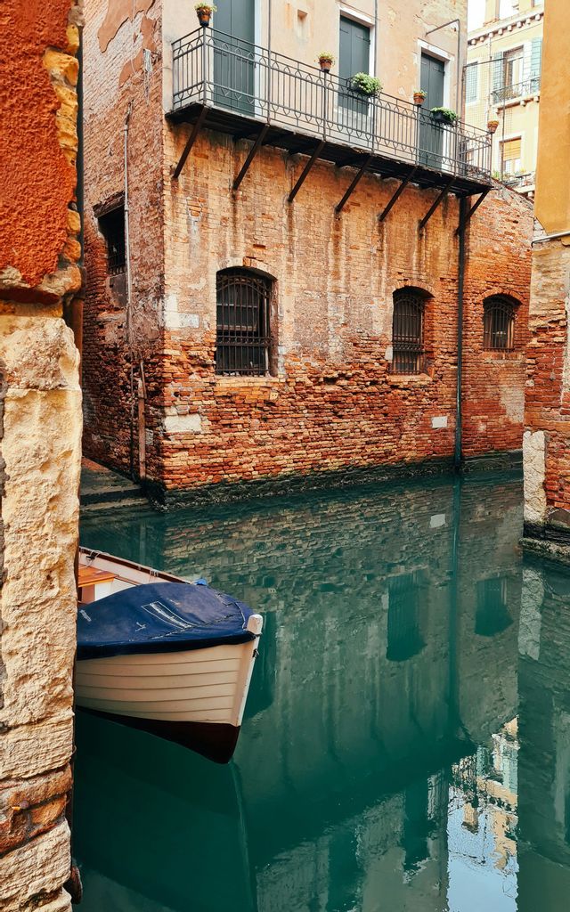 A small boat with a blue cover floats in a narrow canal, its green water reflecting the surrounding weathered brick buildings.