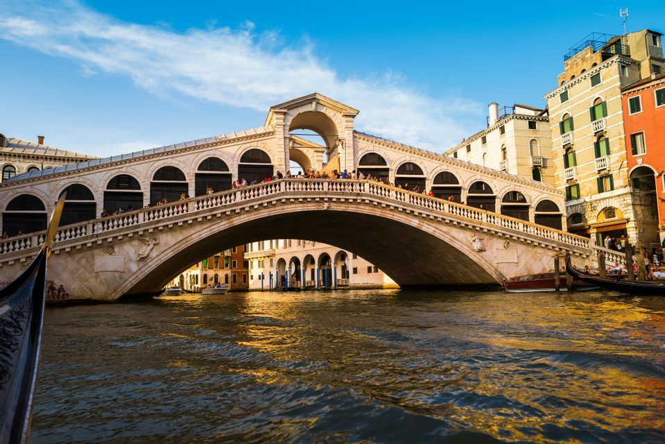 An ornate stone bridge full of people crosses a canal, with golden sunlight reflecting on the water.