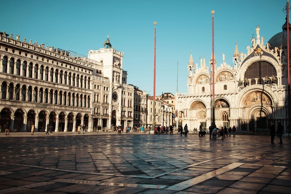 A sunlit historic square with people walking across the wet stone pavement, flanked by an ornate basilica and an arched building.