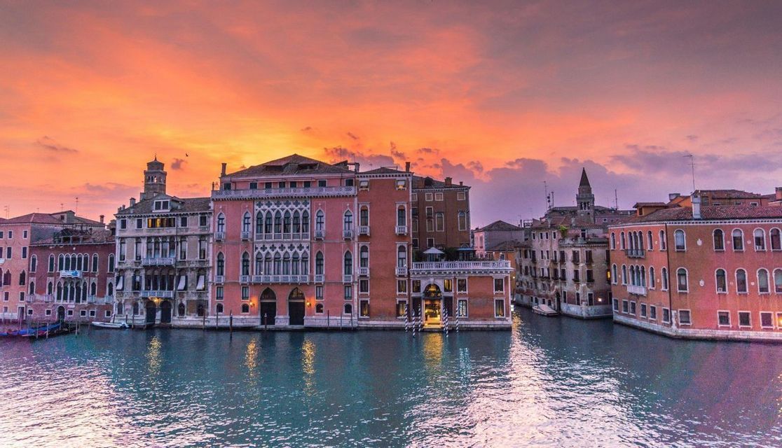 Historic buildings with ornate architecture line a canal under a vibrant orange and pink sunset sky, with reflections in the water.