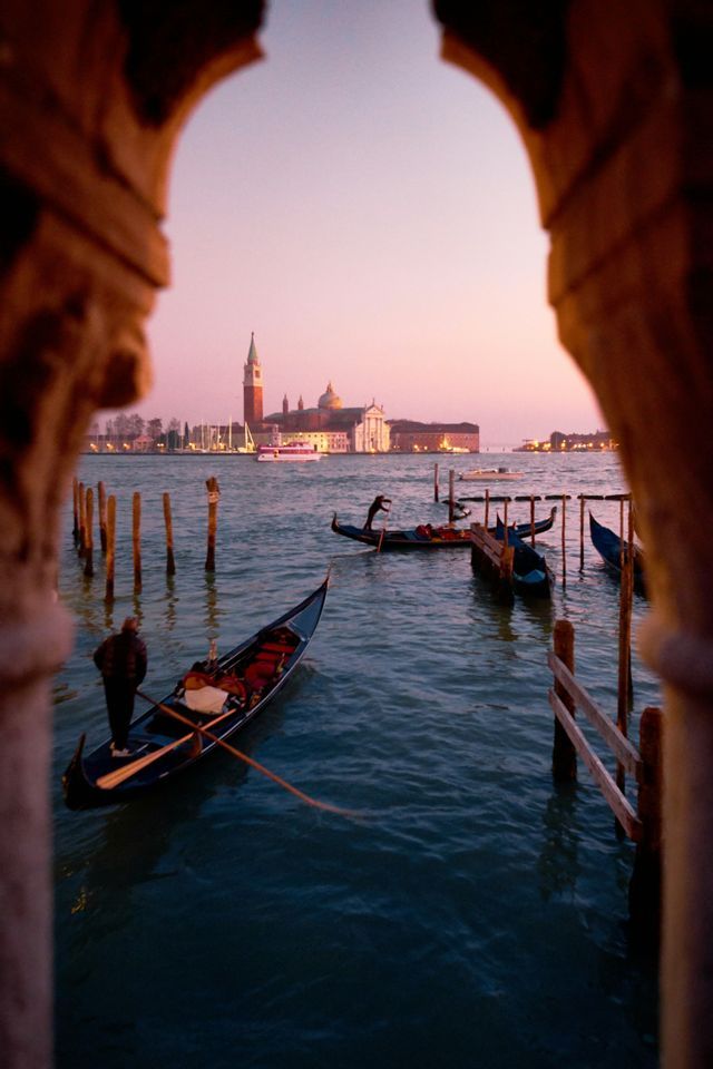 Gondolas float on a canal at dusk, viewed through a stone archway with a distant cityscape on the horizon.