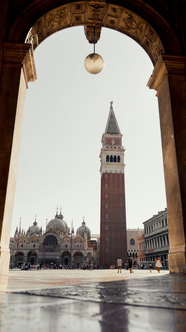 A low-angle view of a red brick bell tower and a domed basilica, framed by a stone archway in a crowded plaza.