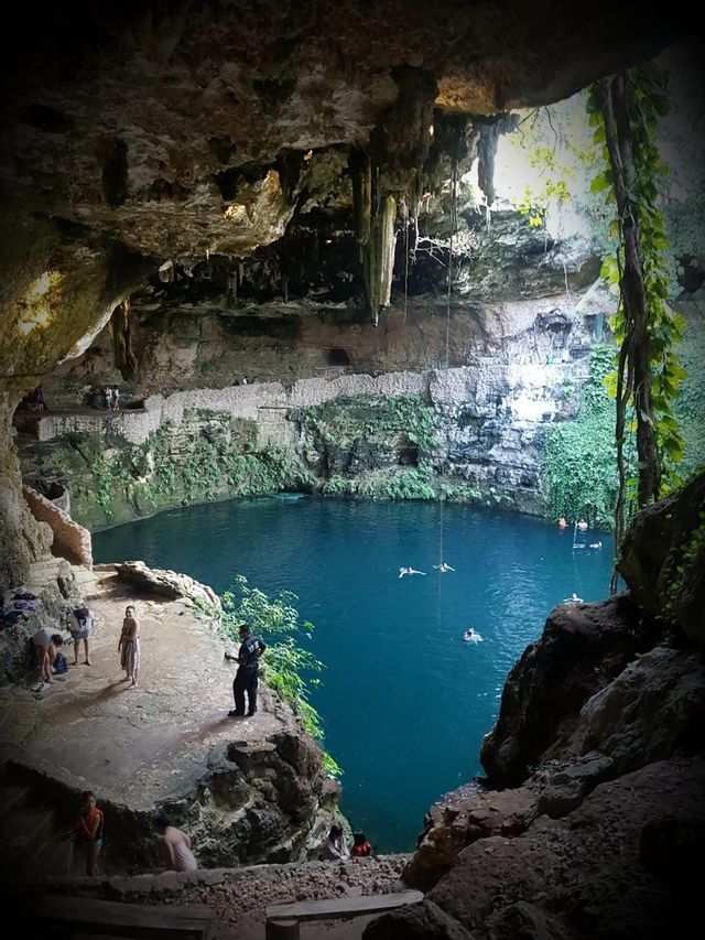 Menschen schwimmen in einem großen, blauen Naturpool in einer sonnenbeschienenen Höhle mit felsigen Felsvorsprüngen und hängenden Tropfsteinen.