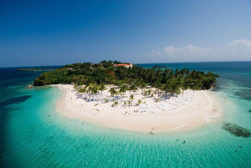 An aerial view of a small tropical island with a white sand beach, numerous palm trees, and clear turquoise water under a blue sky.