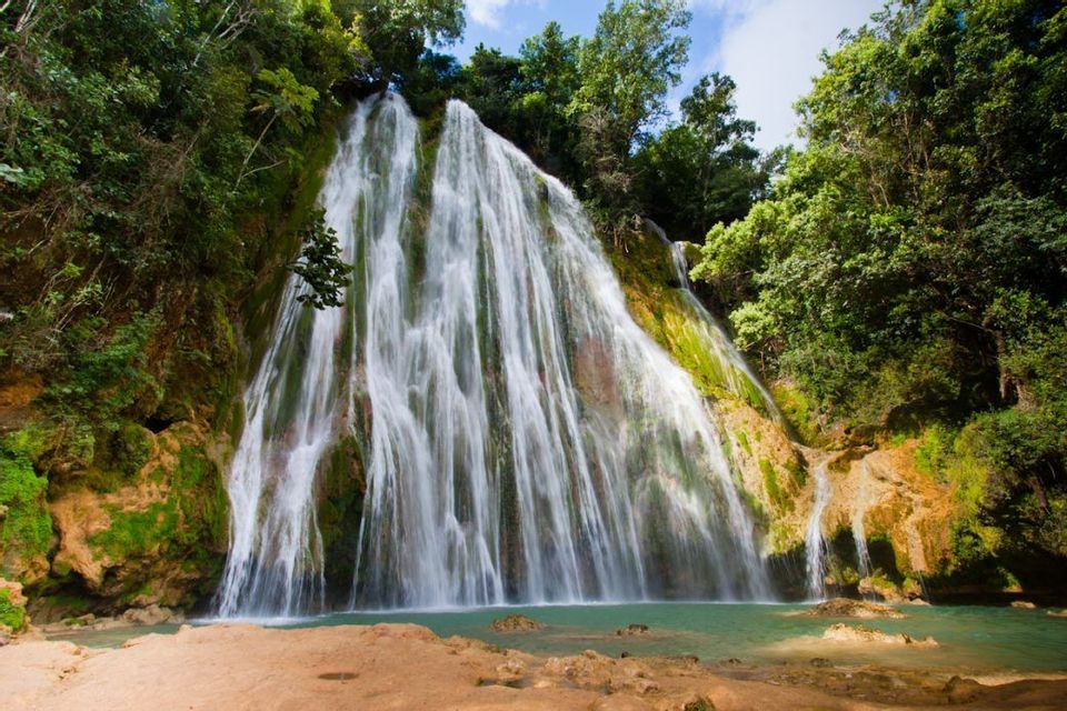 A wide waterfall cascades down a mossy rock face into a turquoise pool, surrounded by dense jungle foliage.